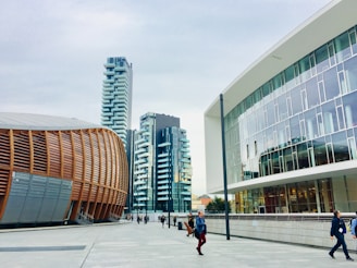 A modern urban setting featuring contemporary architectural designs. On the left is a distinctive building with curved, wooden paneling, suggesting an auditorium or cultural center. Adjacent high-rise buildings with balconies boast a sleek, residential look. The right side showcases a modern office building with large glass windows. Pedestrians walk along the spacious, clean concrete walkway under a cloudy sky.