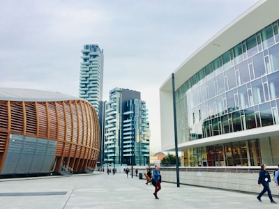 A modern urban setting featuring contemporary architectural designs. On the left is a distinctive building with curved, wooden paneling, suggesting an auditorium or cultural center. Adjacent high-rise buildings with balconies boast a sleek, residential look. The right side showcases a modern office building with large glass windows. Pedestrians walk along the spacious, clean concrete walkway under a cloudy sky.