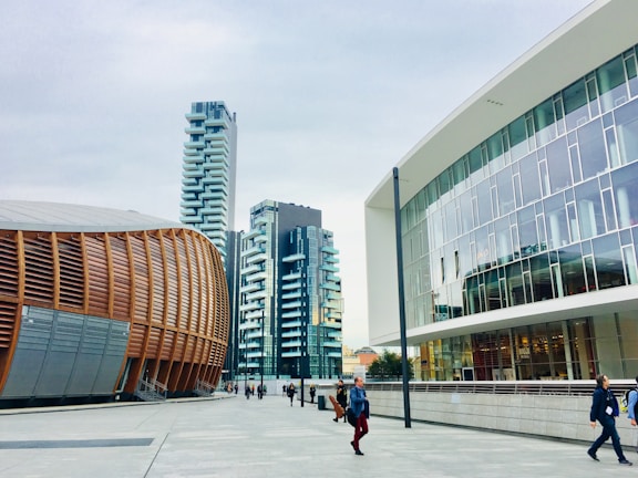 A modern urban setting featuring contemporary architectural designs. On the left is a distinctive building with curved, wooden paneling, suggesting an auditorium or cultural center. Adjacent high-rise buildings with balconies boast a sleek, residential look. The right side showcases a modern office building with large glass windows. Pedestrians walk along the spacious, clean concrete walkway under a cloudy sky.