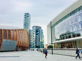 A modern urban setting featuring contemporary architectural designs. On the left is a distinctive building with curved, wooden paneling, suggesting an auditorium or cultural center. Adjacent high-rise buildings with balconies boast a sleek, residential look. The right side showcases a modern office building with large glass windows. Pedestrians walk along the spacious, clean concrete walkway under a cloudy sky.