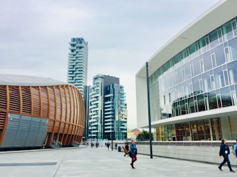 A modern urban setting featuring contemporary architectural designs. On the left is a distinctive building with curved, wooden paneling, suggesting an auditorium or cultural center. Adjacent high-rise buildings with balconies boast a sleek, residential look. The right side showcases a modern office building with large glass windows. Pedestrians walk along the spacious, clean concrete walkway under a cloudy sky.