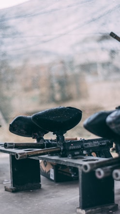 A close-up view of paintball guns placed on a makeshift bench, with the background featuring a blurry view of a netted area. The scene suggests an outdoor, recreational setting, likely at a paintball arena.