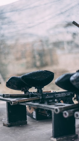 A close-up view of paintball guns placed on a makeshift bench, with the background featuring a blurry view of a netted area. The scene suggests an outdoor, recreational setting, likely at a paintball arena.