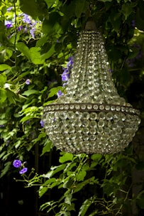 A crystal chandelier is hanging amidst lush green foliage with purple flowers entwined around it. The chandelier's intricate crystal design catches light, adding a touch of elegance to the natural setting.