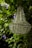 A crystal chandelier is hanging amidst lush green foliage with purple flowers entwined around it. The chandelier's intricate crystal design catches light, adding a touch of elegance to the natural setting.