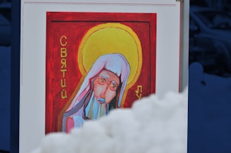 A painting on a display board features a religious figure with a halo, wearing a white headdress and a blue-grey beard. Cyrillic script is visible on either side of the figure. The background is red, and the scene is partially obscured by a mound of snow in the foreground.