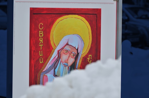 A painting on a display board features a religious figure with a halo, wearing a white headdress and a blue-grey beard. Cyrillic script is visible on either side of the figure. The background is red, and the scene is partially obscured by a mound of snow in the foreground.