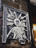 A monochrome sculpture mounted on a brick wall depicts a central figure surrounded by multiple hands holding various objects like cameras, smartphones, guns, and other electronic devices, aimed at the figure. The artwork is framed and shows signs of wear.