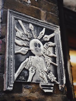 A monochrome sculpture mounted on a brick wall depicts a central figure surrounded by multiple hands holding various objects like cameras, smartphones, guns, and other electronic devices, aimed at the figure. The artwork is framed and shows signs of wear.