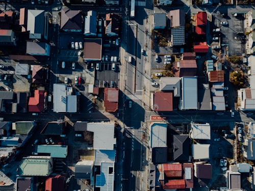 The aerial view showcases a cityscape with a grid-like pattern of streets and buildings. The rooftops vary in color, including shades of red, blue, and gray. Cars are parked in several lots, and the shadows indicate a clear day with the sunlight coming from a low angle.