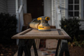 A yellow and black electric sander rests on a wooden cabinet door placed on a pair of sawhorses. The background features a blurred house exterior with a plant, conveying a home improvement setting.