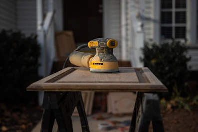 A yellow and black electric sander rests on a wooden cabinet door placed on a pair of sawhorses. The background features a blurred house exterior with a plant, conveying a home improvement setting.