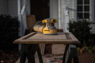 A yellow and black electric sander rests on a wooden cabinet door placed on a pair of sawhorses. The background features a blurred house exterior with a plant, conveying a home improvement setting.