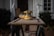 A yellow and black electric sander rests on a wooden cabinet door placed on a pair of sawhorses. The background features a blurred house exterior with a plant, conveying a home improvement setting.