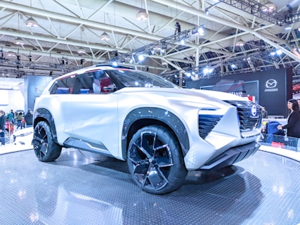 A futuristic white SUV concept car with large black tires is displayed on a platform in an indoor exhibition hall. The vehicle has sleek, angular design elements, prominent side mirrors, and a front grille with a distinctive pattern. Overhead lighting highlights its shiny surface. Several people are visible in the background, observing the car and the exhibition stand.