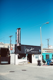 A street scene featuring a small liquor and market store with a prominent sign displaying 'Nick's' above the entrance. The store is situated on a corner, with several signs advertising groceries, wine, and spirits. A lamp post and a utility pole with power lines are visible, along with a few parked cars and trash bins.