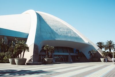 A large, futuristic building with a distinct curved, white dome structure and extensive concrete work. Palm trees in large pots are positioned near the entrance, which features glass doors. The foreground consists of a wide, paved area with clean lines.
