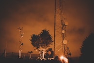 A tall communications tower stands against a cloudy, orange-tinted sky. Several satellite dishes and antennas are attached to the tower. Nearby are smaller structures and a leafy tree, all illuminated by warm, artificial lighting.