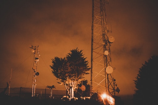 A tall communications tower stands against a cloudy, orange-tinted sky. Several satellite dishes and antennas are attached to the tower. Nearby are smaller structures and a leafy tree, all illuminated by warm, artificial lighting.