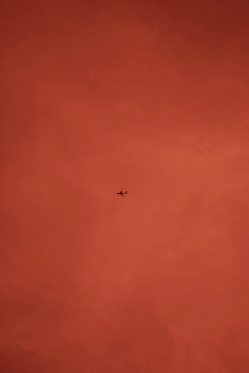 The image depicts a small airplane flying high above an orange sky, which is predominantly red with some clouds scattered throughout. The airplane is positioned centrally within the frame and appears to be moving from left to right across the image.