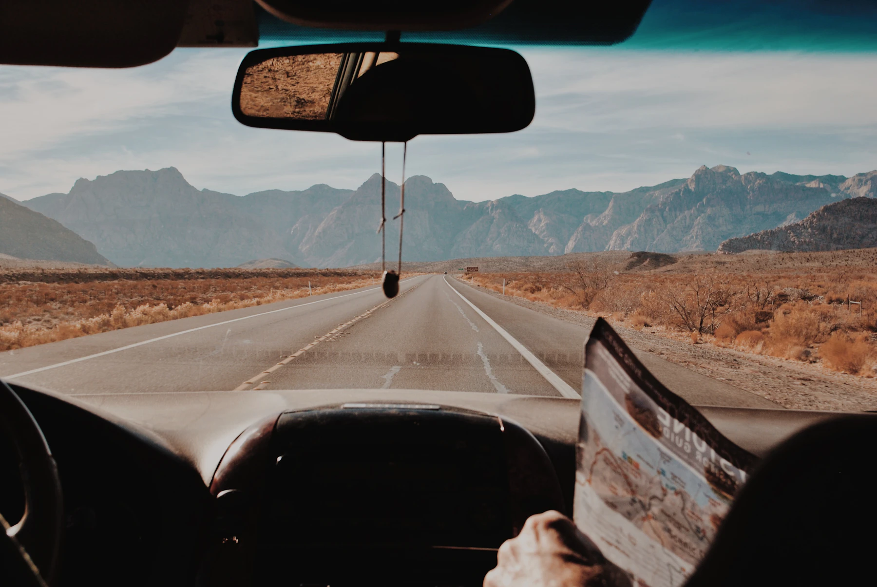 Seen from the back seat, a passenger holding a folded paper map as a two-lane desert road runs toward distant mountains through the windshield