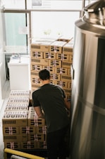 A person wearing a dark shirt is stacking or organizing cardboard boxes labeled 'BROUHAHA' in a room that appears to be a storage area or a brewery. The boxes are stacked in a neat arrangement, and there is a large metallic tank on the right side of the image.
