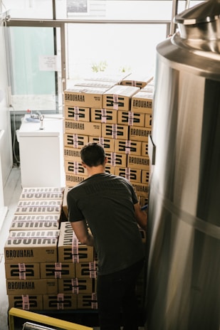 A person wearing a dark shirt is stacking or organizing cardboard boxes labeled 'BROUHAHA' in a room that appears to be a storage area or a brewery. The boxes are stacked in a neat arrangement, and there is a large metallic tank on the right side of the image.