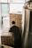A person wearing a dark shirt is stacking or organizing cardboard boxes labeled 'BROUHAHA' in a room that appears to be a storage area or a brewery. The boxes are stacked in a neat arrangement, and there is a large metallic tank on the right side of the image.