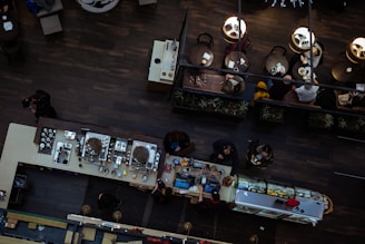 An overhead view of a modern café interior, featuring a long counter with various kitchen equipment and people seated at round tables. The café has dark wood flooring and soft, warm lighting. There are several people seated at the tables, some engaged in conversation, while others are eating.