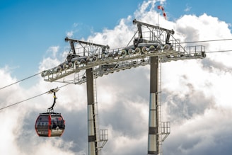 A cable car suspended on a high-altitude lift system with cloudy skies in the background, suggesting a mountain environment. The metal structure supporting the cable car is prominently visible, along with a red wind sleeve that indicates wind direction atop the structure.