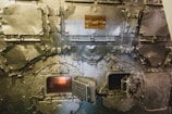 A close-up view of the front of an industrial furnace or boiler, showing metal doors and latches. The surfaces are worn and metallic, with a dull sheen. Two small openings reveal glowing fire inside. A plaque with writing is affixed to the upper part of the machinery.