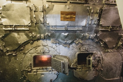 A close-up view of the front of an industrial furnace or boiler, showing metal doors and latches. The surfaces are worn and metallic, with a dull sheen. Two small openings reveal glowing fire inside. A plaque with writing is affixed to the upper part of the machinery.