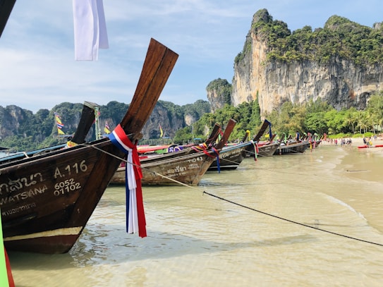 A series of traditional wooden boats with colorful ribbons are docked along a sandy beach. Towering limestone cliffs covered in green vegetation rise in the background. Several people walk along the shoreline, and the sea is calm and clear.