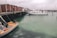 A dock area with several small boats moored in calm, greenish water. The dock has red wooden sheds lined up along it, and a cloudy sky looms overhead, reflecting in the water.