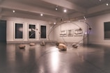 A contemporary art gallery space features an installation comprising large metallic loops intersecting and intertwining with each other. Rocks are strategically placed on and around the loops, creating a dynamic and balanced composition. The walls display abstract monochrome artwork in large frames.