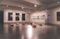 A contemporary art gallery space features an installation comprising large metallic loops intersecting and intertwining with each other. Rocks are strategically placed on and around the loops, creating a dynamic and balanced composition. The walls display abstract monochrome artwork in large frames.
