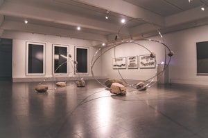 A contemporary art gallery space features an installation comprising large metallic loops intersecting and intertwining with each other. Rocks are strategically placed on and around the loops, creating a dynamic and balanced composition. The walls display abstract monochrome artwork in large frames.