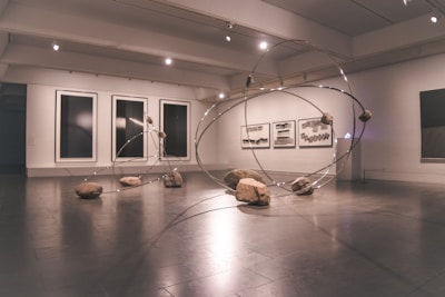 A contemporary art gallery space features an installation comprising large metallic loops intersecting and intertwining with each other. Rocks are strategically placed on and around the loops, creating a dynamic and balanced composition. The walls display abstract monochrome artwork in large frames.