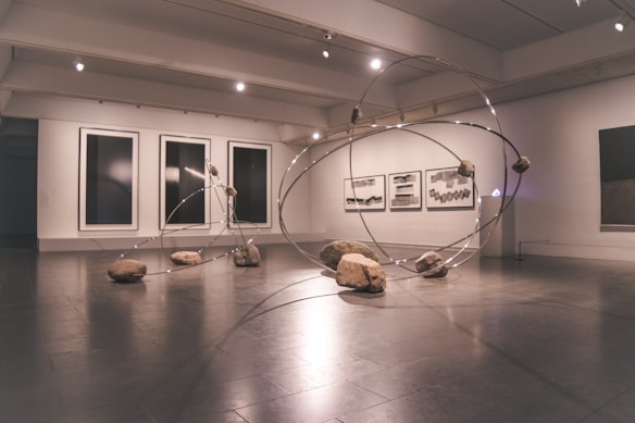A contemporary art gallery space features an installation comprising large metallic loops intersecting and intertwining with each other. Rocks are strategically placed on and around the loops, creating a dynamic and balanced composition. The walls display abstract monochrome artwork in large frames.