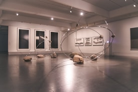A contemporary art gallery space features an installation comprising large metallic loops intersecting and intertwining with each other. Rocks are strategically placed on and around the loops, creating a dynamic and balanced composition. The walls display abstract monochrome artwork in large frames.