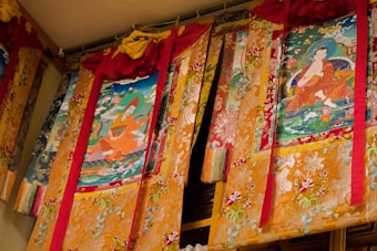 A series of ornate tapestries hang on a wall, featuring colorful and detailed Buddhist artwork. Two primary images depict seated figures in flowing robes surrounded by vibrant flora and abstract patterns. The tapestries are predominantly gold with intricate embroidery, accented by red and pink fabric at the top.