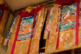 A series of ornate tapestries hang on a wall, featuring colorful and detailed Buddhist artwork. Two primary images depict seated figures in flowing robes surrounded by vibrant flora and abstract patterns. The tapestries are predominantly gold with intricate embroidery, accented by red and pink fabric at the top.