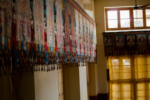 A series of colorful, intricately designed textile banners or tapestries hang along a wall in a traditional interior space. The room is lit by large windows with soft lighting coming through yellow curtains, casting a warm glow.