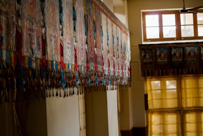 A series of colorful, intricately designed textile banners or tapestries hang along a wall in a traditional interior space. The room is lit by large windows with soft lighting coming through yellow curtains, casting a warm glow.