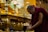 A monk wearing a traditional maroon robe appears to be engaged in a religious or spiritual activity beside an ornate, golden altar. The altar has intricate decorations and a glass case containing a small, detailed structure resembling a temple. The setting is richly adorned with symbolic items, conveying a sacred atmosphere.