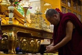 A monk wearing a traditional maroon robe appears to be engaged in a religious or spiritual activity beside an ornate, golden altar. The altar has intricate decorations and a glass case containing a small, detailed structure resembling a temple. The setting is richly adorned with symbolic items, conveying a sacred atmosphere.