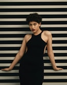 A person with short curly hair is posing against a background of horizontal black and white stripes, wearing a sleeveless black dress.