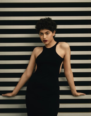 A person with short curly hair is posing against a background of horizontal black and white stripes, wearing a sleeveless black dress.