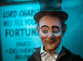 A vintage fortune-teller automaton with a smiling expression, wide-open blue eyes, and rosy cheeks. It wears a black top hat, a black suit, and a white shirt beneath. Behind it, a blurred sign reads 'Lord Charles will tell your fortune,' creating a mystical atmosphere with blue lighting.