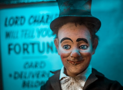A vintage fortune-teller automaton with a smiling expression, wide-open blue eyes, and rosy cheeks. It wears a black top hat, a black suit, and a white shirt beneath. Behind it, a blurred sign reads 'Lord Charles will tell your fortune,' creating a mystical atmosphere with blue lighting.